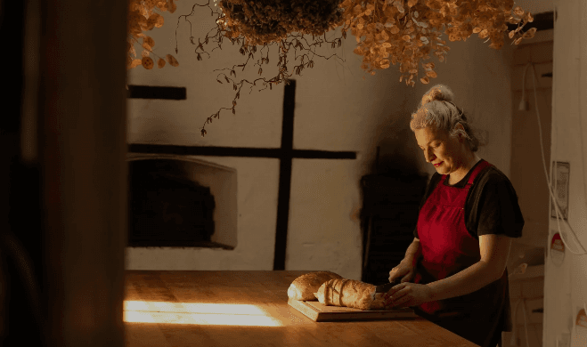 A Cherry Bomb Cafe team member in a cherry-red apron slicing warm bread with dried florals hanging overhead